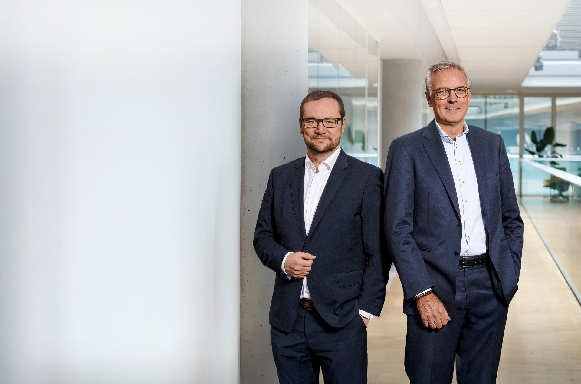 Jens and Claus are standing in a modern, well-lit office environment. Both are wearing dark blue suits with white shirts, conveying a professional, corporate atmosphere. The background features glass windows and doors, reflecting light and showcasing a few plants.